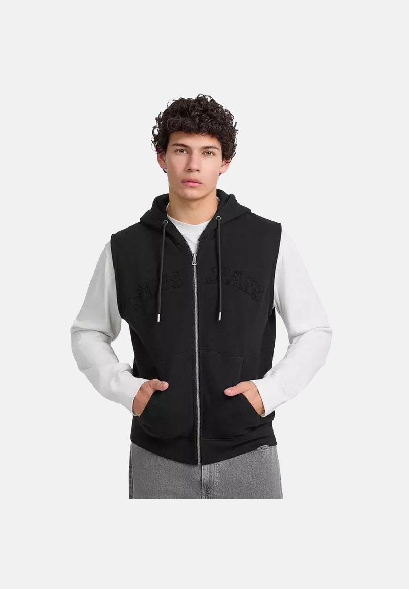 Chaleco negro con cremallera y capucha, con bolsillos delanteros, hecho de una tela suave y llevado sobre una camisa de manga larga blanca.