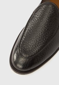 Μαύρο δερμάτινο παπούτσι τύπου slip-on με υφή στο επάνω μέρος, στρογγυλεμένη μύτη και λείο, αντίθετο πέλμα. Λεπτομέρεια ραφής πάνω από την  περιοχή της μύτης.