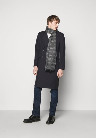 Overcoat blu navy con design doppiopetto, abbinato a una sciarpa grigia a fantasia, jeans scuri e scarpe nere stringate. Accenti in tessuto testurizzato.