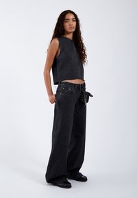 Vrouw met lang, krullend haar, gekleed in een mouwloos cropped zwart denimtop en wijde zwarte denim jeans, staand op een witte achtergrond.