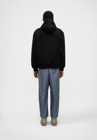 Črna kapuča s teksturiranim zaključkom, kombinirana z svetlo modrimi širokimi hlačami in bež čevlji. Pogled je od zadaj.