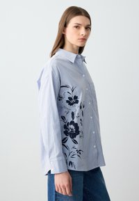 Camicia a maniche lunghe a righe blu e bianche con una stampa floreale nera su un lato; presenta un colletto e una singola tasca sul petto.