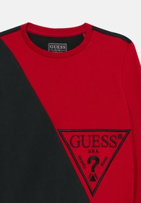 Sudadera roja y negra con cuello redondo. Presenta un gran logo triangular negro bordado con la palabra "GUESS" en la parte frontal.