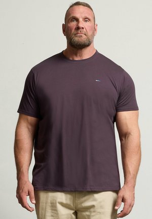 Mężczyzna o muskularnej sylwetce, z krótkimi włosami i brodą, ubrany w jednolitą ciemnofioletową koszulkę i beżowe spodnie, stojący na neutralnym tle.