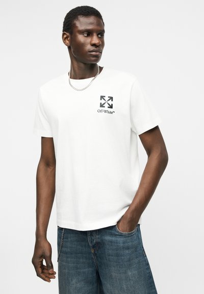 Άνδρας που φοράει λευκό μπλουζάκι Off-White με μαύρο λογότυπο στο στήθος, μπλε τζιν, ασημένιο κολιέ αλυσίδα, στέκεται με το ένα χέρι στην τσέπη, κοιτώντας προς τα πλάγια.