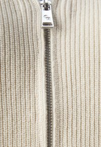 Beige Strickpullover mit metallischem Reißverschluss. Besitzt eine gerippte Textur und auffällige Nähte an der Vorderseite.