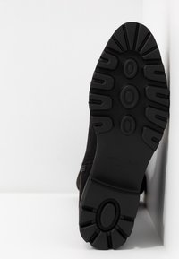 Semelle d'une botte noire avec des crampons profonds, des motifs ovales et une adhérence texturée, positionnée verticalement contre un mur et un sol blancs.