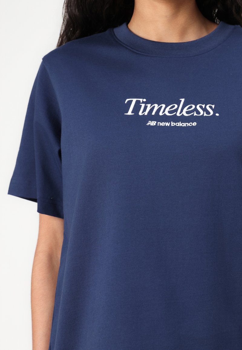 T-shirt en coton bleu marine à manches courtes, avec un logo brodé blanc indiquant "Timeless." et "new balance" en dessous. Design simple.