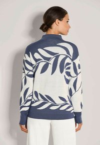 Frau mit dunklem Haar in einem tiefen Dutt, die einen blau-weißen Pullover mit einem großen Blättermuster trägt, kombiniert mit weißen Hosen, von hinten blickend.