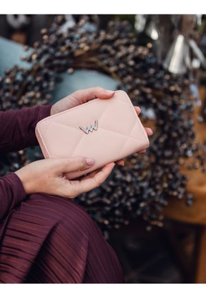 Pink quilted wallet with silver logo, rectangular shape, textured surface, held in hands, with a blurred background of decorative elements.