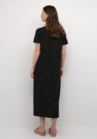 Black cotton maxi dress with short sleeves, straight cut, and ankle length. Smooth texture and simple design. Worn with flat, woven sandals.