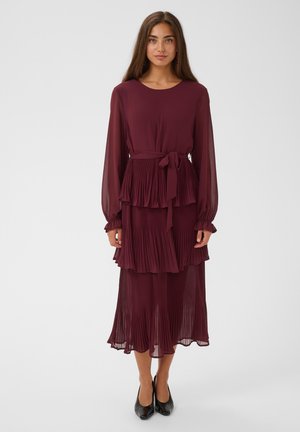 Mujer con un vestido burdeos de manga larga con capas plisadas y un cinturón atado en la cintura, combinado con zapatos negros de tacón, de pie sobre un fondo blanco.
