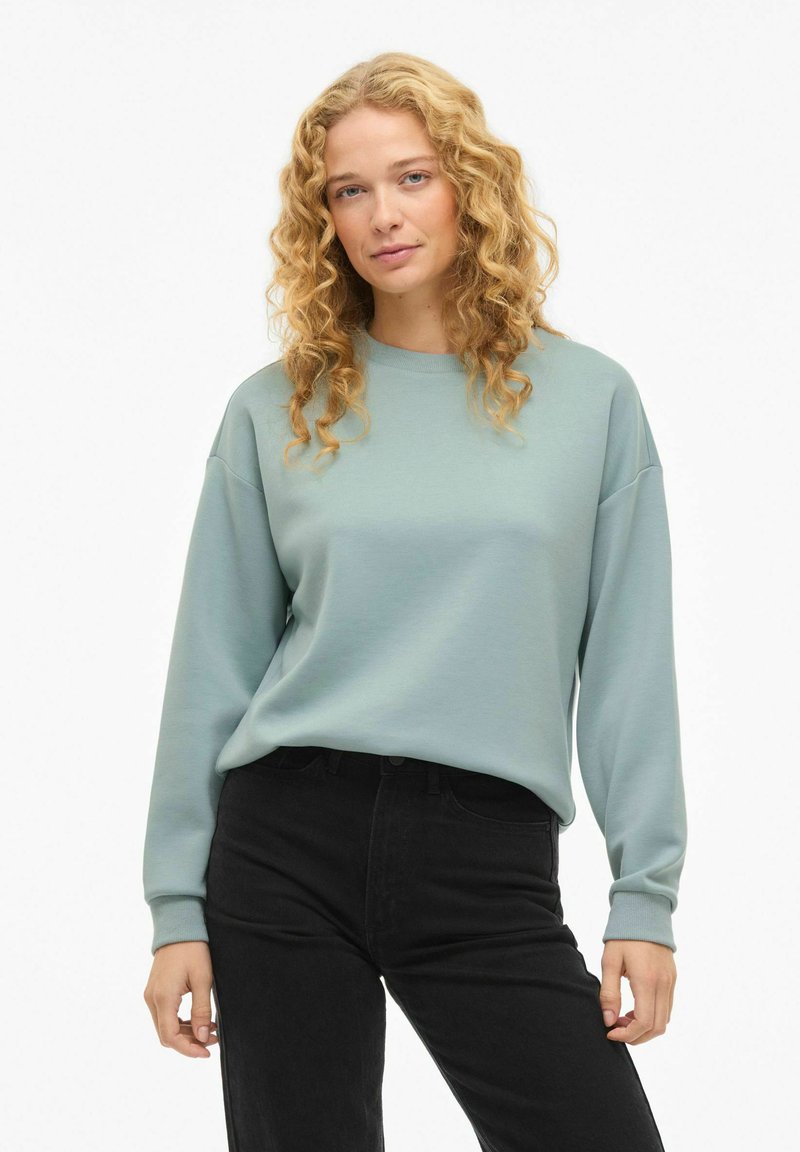 Svetlo zelena majica z okroglim ovratnikom in spuščenimi rameni, združena s črnimi kavbojkami. Mehka tkanina, sproščen kroj, brez vidnih vzorcev.