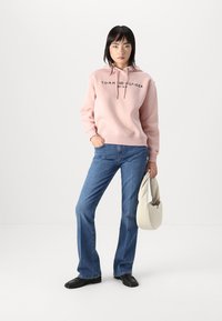 Haut à capuche rose clair avec logo noir, associé à un jean évasé bleu. Des chaussures sombres et un sac à main beige clair complètent la tenue.