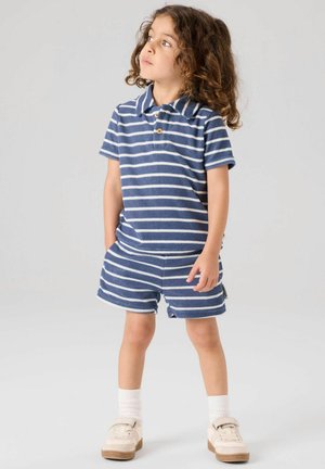 Niño con cabello rizado que lleva una camiseta de manga corta y pantalones cortos a rayas azules y blancas, de pie con las manos en los bolsillos.