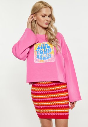 Blond kvinde iført lyserød sweatshirt med teksten "Lev din drøm" og farverigt stribet nederdel, rører ved håret og kigger ned.
