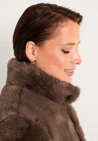 Brun faux fur frakke med høj krave, der har en glat overflade og synlige sømme. Gyldne, sfæriske øreringe tilføjer detaljer.
