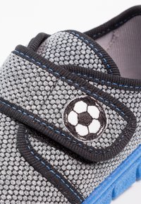 Szare buty z wzorzystego materiału z czarnymi akcentami i niebieskimi szwami, z naszywką w kształcie piłki nożnej na pasku oraz teksturowaną podeszwą.