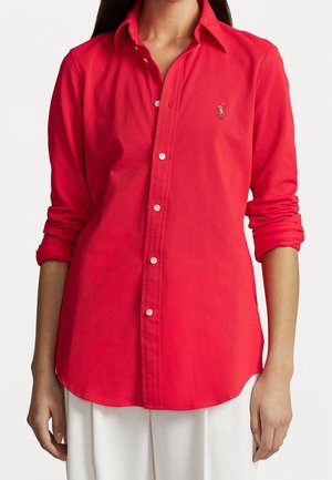 Vrouw draagt een felrode blouse met opgekrulde mouwen en een geborduurd logo op de linkerborst, gecombineerd met witte broek.