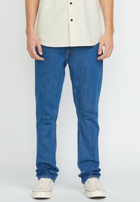Jean en denim bleu à coupe ajustée, présentant un design classique avec une texture lisse. Porté avec une chemise boutonnée rayée légère et des baskets blanches.