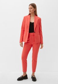 Koraalkleurig pak met een getailleerde blazer met gepunte revers en bijpassende, op maat gemaakte broeken. Vergezeld van zwarte schoenen met een decoratieve accent.