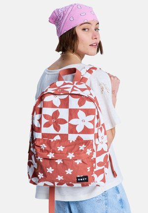 Femme portant une chemise blanche et un bandana rose, avec un sac à dos à motifs floraux rouges et blancs porté sur une épaule.