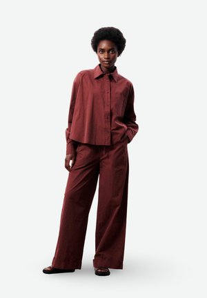 Femme portant une chemise bordeaux ample à boutons et un pantalon à jambes larges, debout avec une main dans la poche, sur fond blanc.