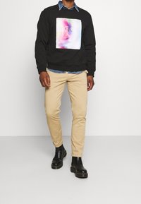 Sudadera negra con un cuadrado gráfico multicolor, pantalones slim-fit beige y botas negras hasta el tobillo. Superpuesta con una camisa de mezclilla por debajo.