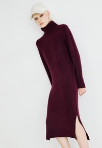 Burgundy knit dress with a high turtleneck, long sleeves, and side slit detail. Soft texture and form-fitting design.
