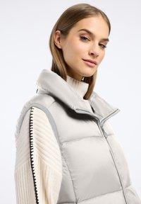 Jasnoszara kamizelka puchowa z wysokim kołnierzem, zapięciem na zamek i beżowym swetrem o teksturowanym splocie na dole. Posiada kontrastowe przeszycia na rękawach.