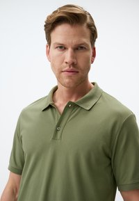 Polo shirt verde oliva realizzato in cotone morbido. Presenta un colletto classico, una patta con un pulsante e maniche corte.