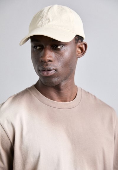 Casquette en coton beige avec des détails brodés, portée avec un t-shirt à col rond marron. Le tissu semble doux et légèrement texturé.
