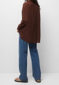 Chemise marron à manches longues, ample, avec un tissu texturé, assortie à un jeans en denim bleu à jambes larges et des tongs décontractées.