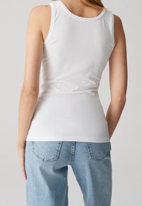 Femme portant un haut blanc moulant sans manches et un jean denim bleu clair, vue de dos sur un fond uni.