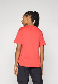Red short-sleeve t-shirt with a crew neckline. Features subtle lettering along the side. Worn with black shorts, background is plain grey.