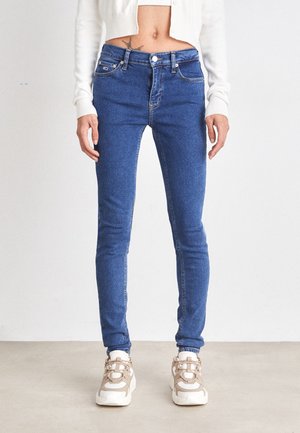 Jean skinny en denim bleu moyen, à taille haute, avec un design à cinq poches et des détails de couture subtils, associés à des baskets beiges.