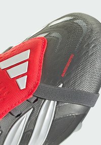 Botín de fútbol negro y rojo con una parte superior texturizada, acentos plateados y correa ajustable. El diseño incluye tres rayas reflectantes.