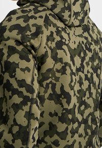 Mikina s kapucí v maskáčovém vzoru tmavě zelen é, olivové a černé barvy. Vyrobena z měkké, elastické tkaniny s viditelnými detaily šití.