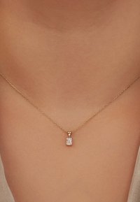 Goldkette mit einer feinen Kette, die einen rechteckigen klaren Edelstein-Anhänger trägt und auf einer glatten Hautoberfläche liegt.