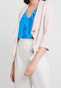 Ljusrosa kavaj med öppna front och trekvartslånga ärmar, matchad med en blå blus med volanger och vita randiga högmidjade byxor.