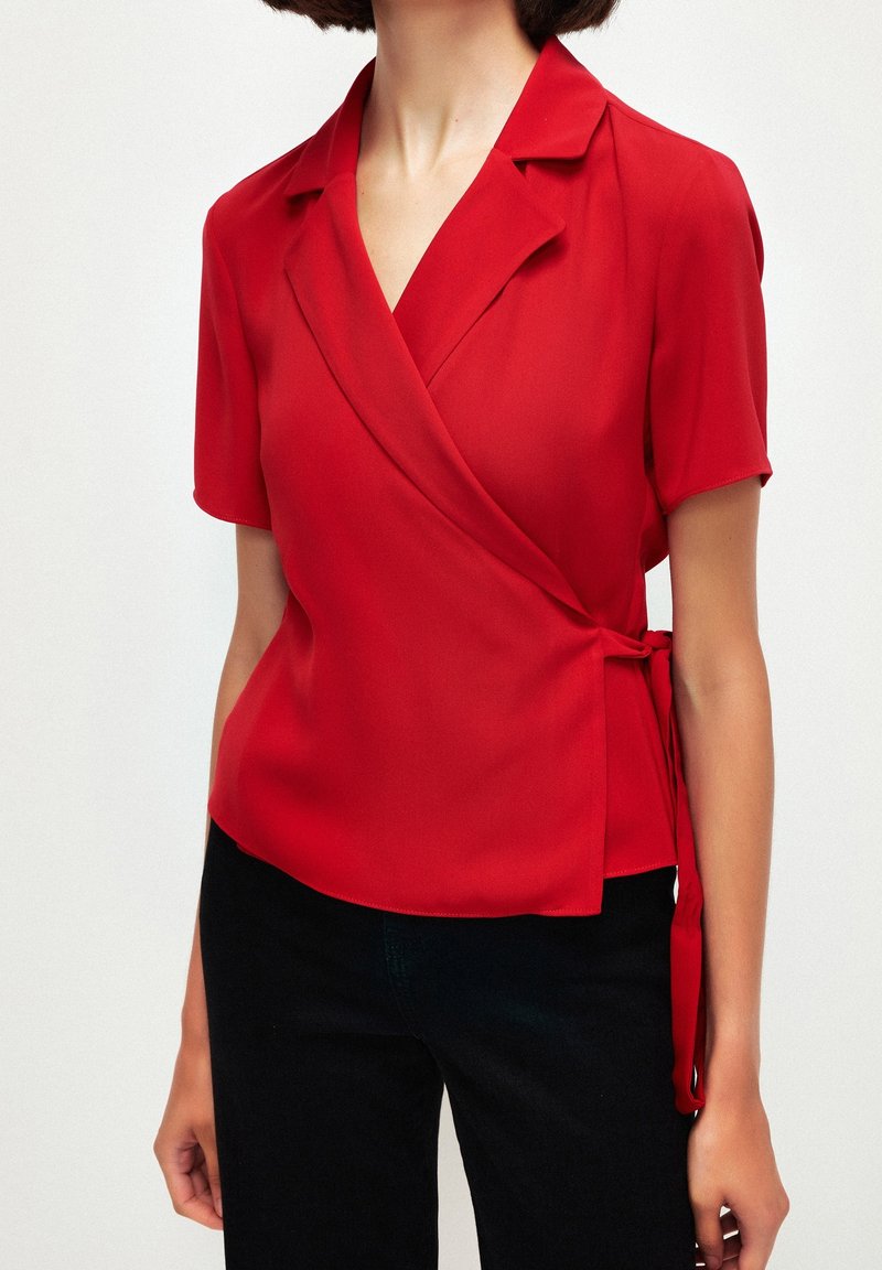 Blouse portefeuille rouge à manches courtes, encolure en V et détail noué sur le côté. Tissu lisse avec une finition légère et élégante.