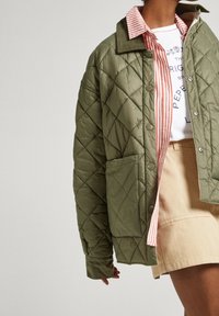 Chaqueta verde acolchada con patrón de diamantes, que cuenta con un cuello, botones delanteros y un bolsillo lateral. Debajo, se ve una camisa a rayas.