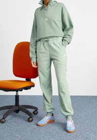 Ljusgrön fleece-träningsdräkt som består av en jacka med dragkedja och byxor med elastisk midja, i kombination med blå sneakers och placerad bredvid en orange stol.