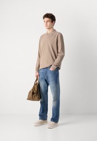 Beige langermet polo, blå jeans, lyse sneakers, holder en brun lærveske. Enkelt design med avslappet passform og uformell stil.