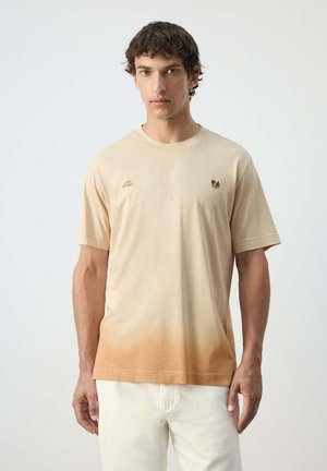 Hombre con cabello corto y oscuro, vistiendo una camiseta de manga corta en tonos beige y marrón claro ombré y pantalones blancos, de pie frente a un fondo liso.