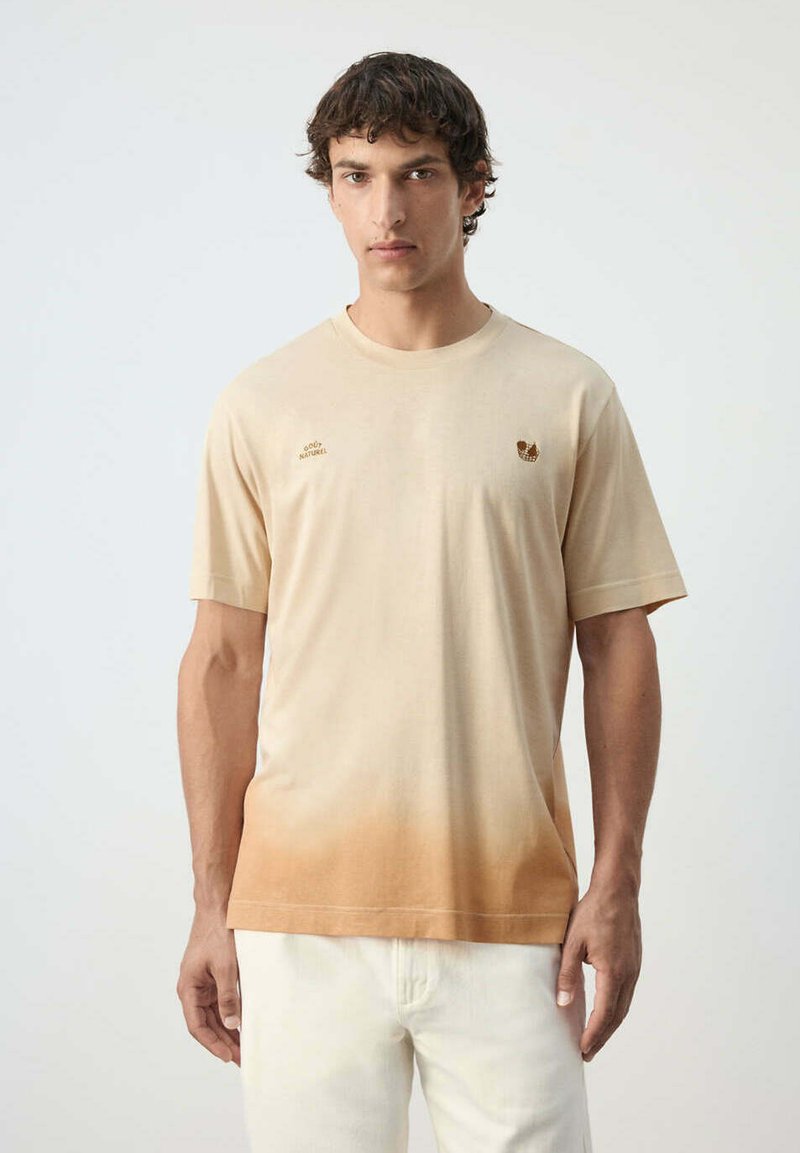Homme aux cheveux courts et foncés portant un t-shirt manches courtes beige et marron clair en dégradé et un pantalon blanc, debout devant un fond uni.