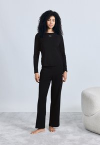 Must pika varrukatega ülemine osa, millel on rinnal "BOSS" logo, sobitatud mustade pükstega, mõlemad tehtud pehmest kangast, hele taust.