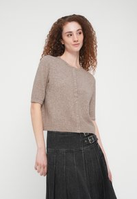 Πλεκτή ελαφρώς καφέ ζακέτα με κοντά μανίκια, στρογγυλή λαιμόκοψη και επτά κουμπιά μπροστά, συνδυασμένη με σκούρα φούστα.