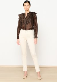 Ruda raukšlių palaidinė su aukštu kaklu, sagų uždarymas, derinama su prigludusiomis grietinėlės spalvos kelnėmis ir béžinėmis kulkšniais ant šviesių medinių grindų.