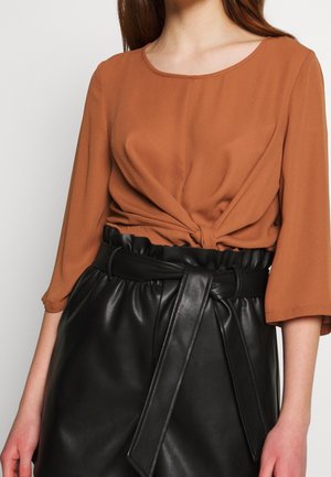 Vrouw met bruine top met gedraaide knoop en driekwartmouwen, ingestopt in een hoogtailles leren zwarte rok met brede riem die aan de voorkant is gestrikt.